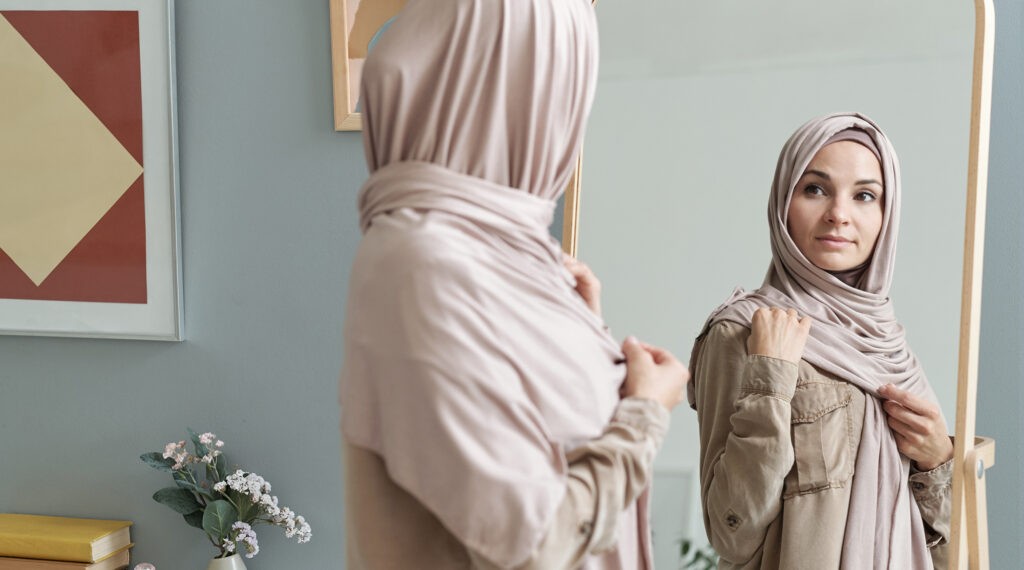 A young woman wearing a light beige hijab stands in front of a mirror, adjusting her hijab while looking at her reflection.