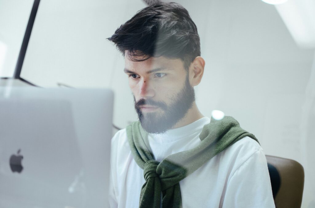 A man with a beard and dark hair intensely focused on his work, looking at a computer screen. He is wearing a white shirt with a green sweater draped over his shoulders, and the setting appears to be a modern, well-lit office.