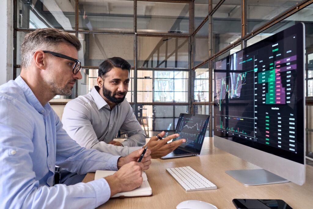 Two men are working together at a desk in a modern office. They are looking at a computer monitor displaying financial charts and data. One man is gesturing toward the screen while the other takes notes in a notebook. Both appear focused and engaged in their work.