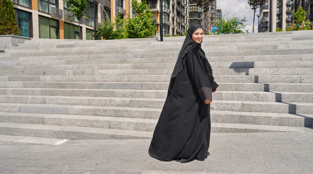 A young woman wearing a black abaya and hijab is walking confidently up a wide set of stone steps in an urban area. She turns to smile at the camera, her expression warm and inviting.