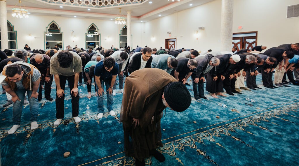 A congregation of worshippers bows in unison during prayer at a mosque, with a religious leader in the front row.