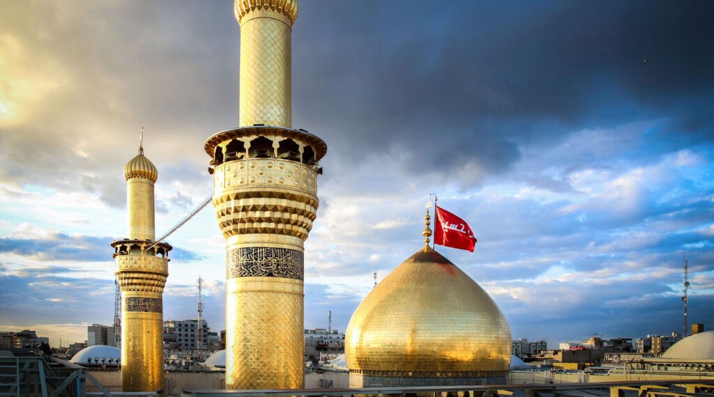 The golden dome and minarets of Imam Hussain's shrine in Karbala, Iraq, with a red flag symbolizing the martyrdom of Imam Hussain (AS) flying high. The background shows a cloudy sky with the cityscape in the distance.