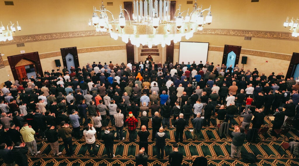 A large congregation of worshippers stands in prayer inside a beautifully ornate mosque. The chandelier above illuminates the space, while Arabic calligraphy decorates the walls.