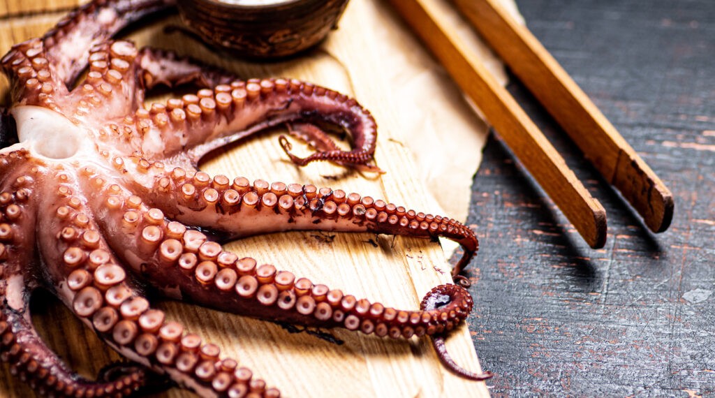 Cooked octopus tentacles laid out on a wooden board next to wooden tongs.