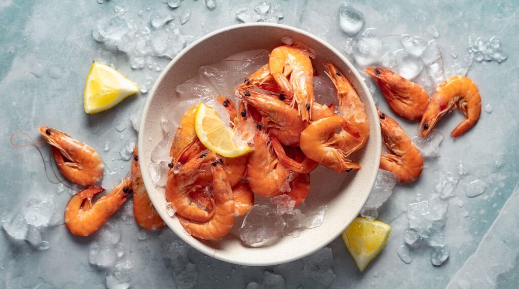 A bowl of cooked shrimp on ice with lemon wedges, surrounded by scattered shrimp and ice cubes.