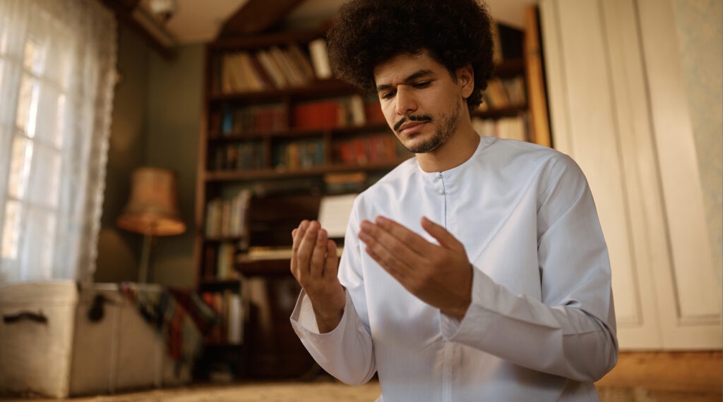 A man wearing traditional white attire, with hands raised in supplication, praying in a room filled with books and natural light.