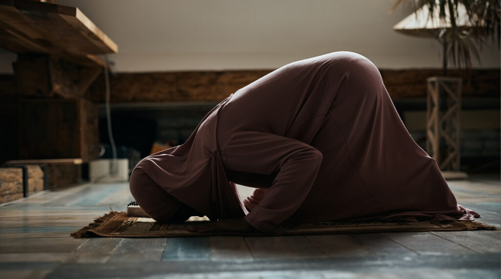 A woman in modest clothing performing sujood (prostration) during prayer in a dimly lit room.