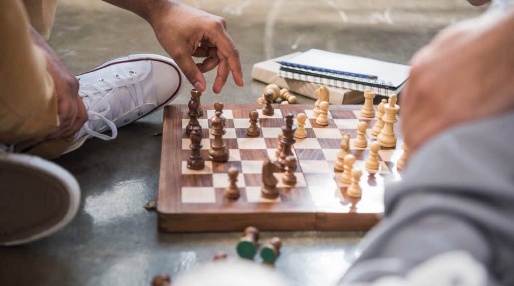  Close-up of a wooden chessboard placed on the ground, with two players visible—one wearing white sneakers and beige pants—strategically moving chess pieces. A notebook lies in the background.