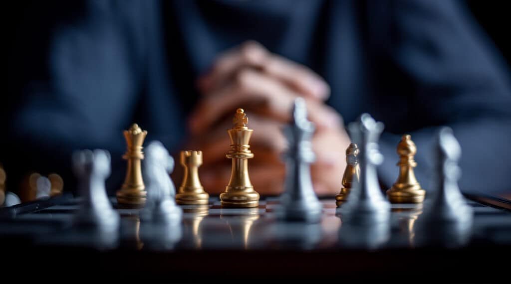 A chessboard with golden and silver chess pieces in focus, positioned against a dark, blurred background. Hands clasped in thought can be seen in the background