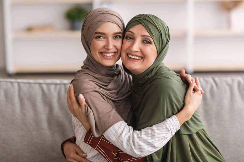 Cheerful Mother And Her Adult Daughter Embracing Posing Wearing Hijab And Smiling To Camera Sitting On Sofa At Home.