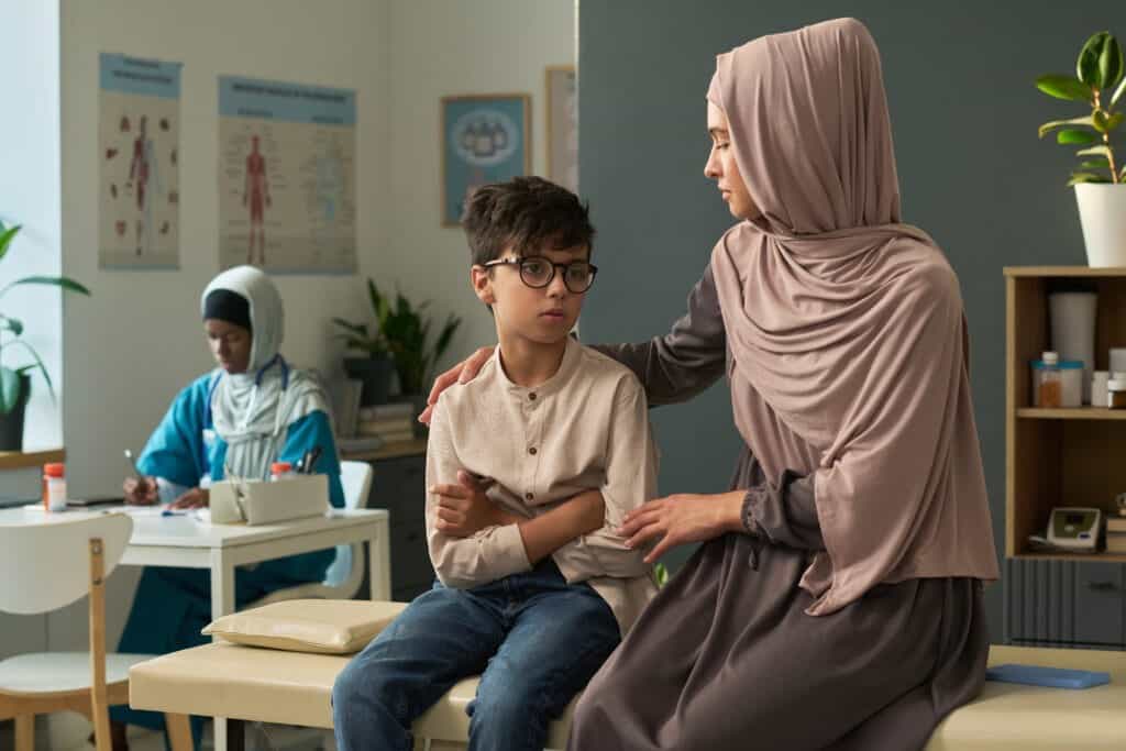 Muslim woman in hijab consoling son during visit to doctor's office while other nurse attending to other patient in background, emphasizing care and comfort