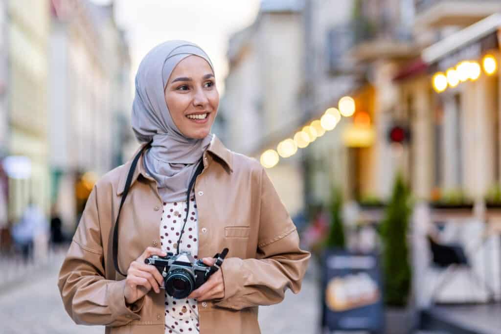 Young woman walking in the evening city in hijab, tourist with camera and wearing a hat inspects the historical city smiling Muslim woman on a trip.