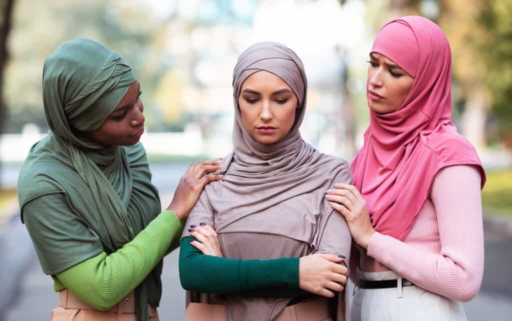 Three Muslim Women Supporting And Comforting Unhappy Female Friend Standing Outdoors.